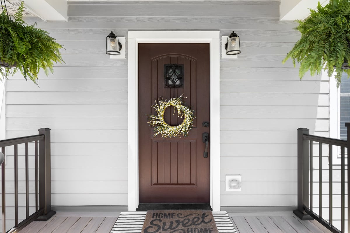 A wooden front door detail with grey vinyl siding, composite decking floor and raining, and lights mounted aside the doorway.