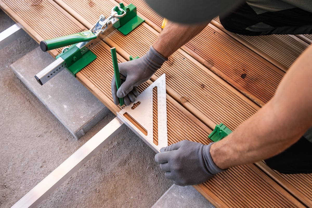 A craftsman carefully places a square tool on wooden planks during a deck renovation outdoors, ensuring precise alignment for a sturdy finish.