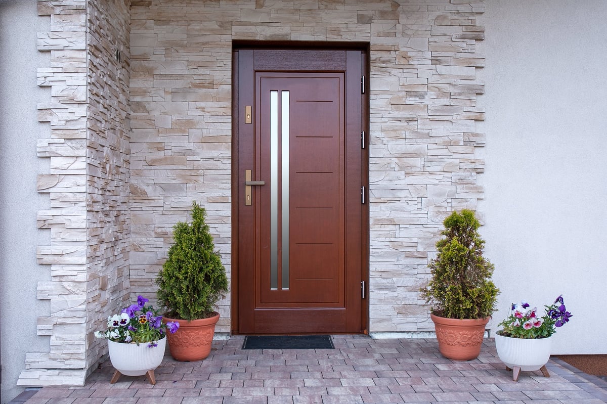 A front door detail with a stone accent wall, a brown door, covered porch, white posts and railing, and white siding.